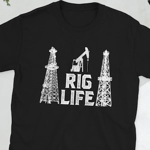 Może przedstawiać: Czarny t-shirt z białym nadrukiem przedstawiającym trzy platformy wiertnicze i napisem "RIG LIFE" poniżej. Projekt przedstawia dwie wieże wiertnicze i pompę naftową. Koszulka wykonana jest z miękkiego, wygodnego materiału.