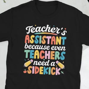 以下が含まれることがあります： カラフルな文字で「Teacher's Assistant because even TEACHERS need a SIDEKICK」と書かれた黒いTシャツ。鉛筆のイラストがデザインされています。楽しくて教育的なテーマのTシャツです。