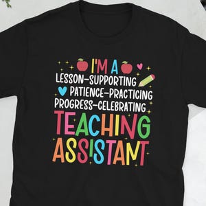 Puede incluir: Camiseta negra con texto colorido que dice "I'M A LESSON-SUPPORTING, PATIENCE-PRACTICING, PROGRESS-CELEBRATING TEACHING ASSISTANT". El diseño incluye gráficos de manzana y lápiz. Un regalo perfecto para un asistente de enseñanza.
