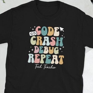Op de afbeelding: Zwart t-shirt met de tekst "CODE CRASH DEBUG REPEAT" in een kleurrijk retro-lettertype. De tekst bevat een codeersymbool en de woorden "Tech Teacher" eronder.