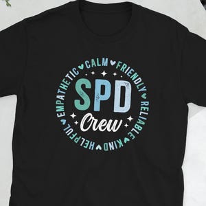 May include: A black short-sleeved t-shirt featuring a circular graphic with the text SPD Crew in the center. Surrounding the text are the words Calm, Friendly, Reliable, Kind, Helpful, and Empathetic in a light blue font with small heart icons.