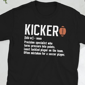May include: Black t-shirt featuring the word "KICKER" in white, with a football graphic. The shirt includes a definition of "KICKER" and their role in the sport. A great gift for football fans.