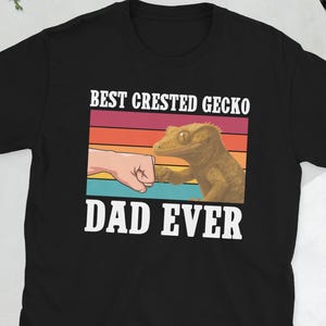 Peut inclure: T-shirt noir avec l'inscription blanche "BEST CRESTED GECKO DAD EVER". Le motif comprend une poignée de main entre une main et un gecko à crête, sur fond de coucher de soleil rétro.