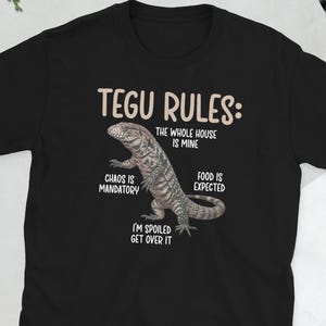 May include: Black t-shirt featuring the text "TEGU RULES" and a lizard graphic. The text below the lizard reads "CHAOS IS MANDATORY", "THE WHOLE HOUSE IS MINE", "FOOD IS EXPECTED", and "I'M SPOILED GET OVER IT".