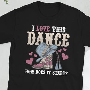 以下が含まれることがあります： 「I LOVE THIS DANCE」の文字と、カウボーイブーツと帽子のイラストが描かれた黒いTシャツ。ピンクのハートが描かれています。「HOW DOES IT START?」の文字が下にあります。