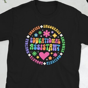May include: Black t-shirt featuring a vibrant circular design. The design includes the words "Educational Assistant" in a retro font, surrounded by flowers, hearts, and words such as "Inspire" and "Encourage".