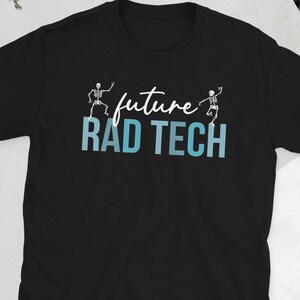 May include: Black t-shirt with the text "future RAD TECH" in white and blue. The design includes two skeleton figures. A camera, glasses, a phone, and a wallet are in the background.