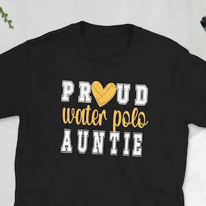 May include: Black t-shirt with the text "PROUD water polo AUNTIE" in white and yellow. The word "PROUD" is in a block letter font, and the word "AUNTIE" is in a similar font. A yellow heart with a water polo ball is in the center.