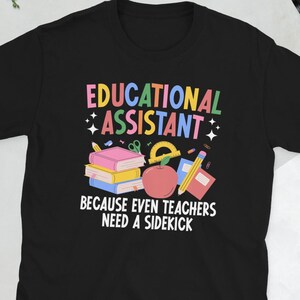 Puede incluir: Camiseta negra con la frase "EDUCATIONAL ASSISTANT" en letras coloridas, junto con un gráfico de libros, una manzana y un lápiz. El texto "BECAUSE EVEN TEACHERS NEED A SIDEKICK" está debajo. La camiseta es de cuello redondo y manga corta.
