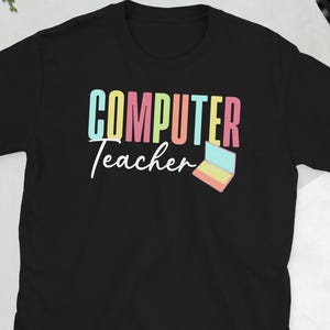 Op de afbeelding: Zwart t-shirt met de tekst "COMPUTER Teacher" in kleurrijke blokletters en een kleine laptopafbeelding. Het lettertype is speels, geschikt voor computerleraren.