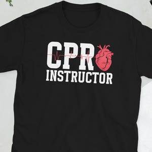 May include: Black t-shirt with the words "CPR INSTRUCTOR" in white block letters. A red heart graphic is to the right of the text. A red heartbeat line is above the letters "CPR".
