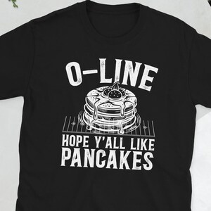 Könnte beinhalten: Schwarzes T-Shirt mit einem weißen Aufdruck von Pfannkuchen und dem Text "O-LINE HOPE Y'ALL LIKE PANCAKES". Das Design zeigt einen Stapel Pfannkuchen mit Sirup. Das T-Shirt ist kurzärmlig.
