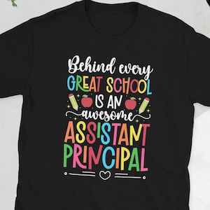 Può includere: Una t-shirt nera a maniche corte con il testo colorato Behind every GREAT SCHOOL IS AN awesome ASSISTANT PRINCIPAL, decorata con illustrazioni di due mele rosse e due matite gialle, oltre a un piccolo simbolo di cuore bianco.