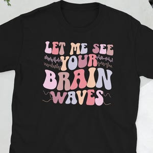 May include: Black t-shirt featuring the phrase "LET ME SEE YOUR BRAIN WAVES" in a retro font. The text is in shades of pink, orange, and purple. The shirt has short sleeves.