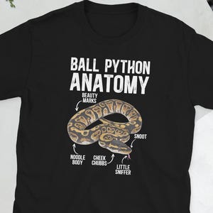 May include: Black t-shirt with a graphic of a ball python anatomy diagram. The diagram includes labels for "Beauty Marks," "Noodle Body," "Cheek Chubbs," "Little Sniffer," and "Snoot." The text "Ball Python Anatomy" is printed above the diagram.