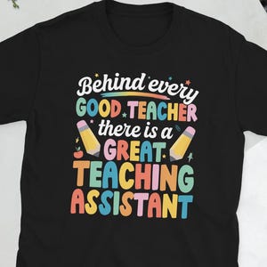 以下が含まれることがあります： カラフルな文字で「Behind every GOOD TEACHER there is a GREAT TEACHING ASSISTANT」と書かれた黒いTシャツ。デザインには2本の鉛筆が含まれています。 ティーチングアシスタントへのプレゼントに最適です。