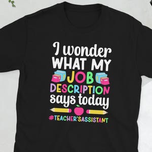 Puede incluir: Camiseta negra con el texto "I wonder what my job description says today" en blanco, rosa, azul y verde. La camiseta también tiene un lápiz y una manzana, y el hashtag #teacher'sassistant.