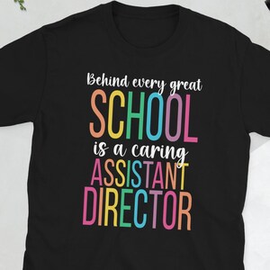 May include: Black t-shirt with the text "Behind every great SCHOOL is a caring ASSISTANT DIRECTOR" in colourful letters. The shirt is a short-sleeved crew neck style.
