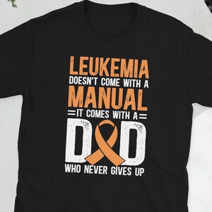 May include: Black t-shirt with the text "LEUKEMIA DOESN'T COME WITH A MANUAL =IT COMES WITH A= D&D WHO NEVER GIVES UP" in orange and white. The design includes an orange awareness ribbon.