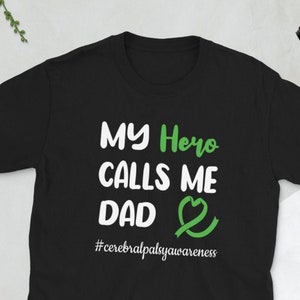 Op de afbeelding: Zwart T-shirt met witte tekst die "My Hero Calls Me Dad" zegt met een groen lint en een hart. De tekst "#cerebralpalsyawareness" staat onder de hoofdtekst.