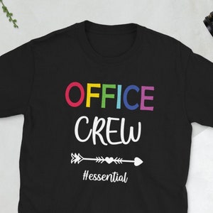Op de afbeelding: Zwart t-shirt met de woorden "OFFICE CREW" in kleurrijke letters. Daaronder een witte pijl met een hartontwerp en het woord "#essential". Een camera, portemonnee, bril en telefoon staan op de achtergrond.