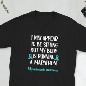 May include: Black t-shirt with white text that reads "I may appear to be sitting but my body is running a marathon #dysautonomia awareness". The text is surrounded by two teal awareness ribbons.