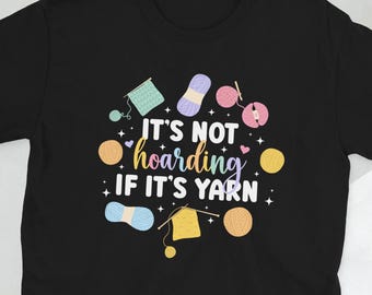 Op de afbeelding: Zwart T-shirt met een kleurrijk grafisch ontwerp met garenbollen, breinaalden en de tekst "Het is geen hamsteren als het garen is".