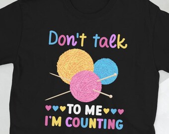 Op de afbeelding: Zwart T-shirt met de tekst "Praat me niet aan, ik ben aan het tellen" in kleurrijke letters. De tekst wordt omringd door drie bollen garen en breinaalden.