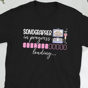 Puede incluir: Camiseta negra con el texto "SONOGRAPHER in progress loading..." en blanco y rosa. El diseño incluye una imagen de ultrasonido en una pantalla de ordenador y una barra de carga rosa y morada.