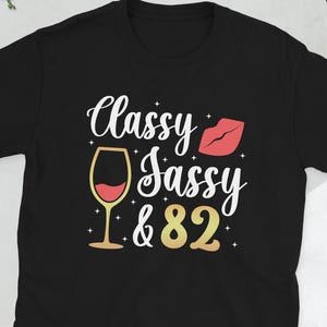 May include: Black t-shirt with the text "Classy Sassy & 82" in white and gold lettering. A red lip print and a glass of wine are also featured on the shirt.
