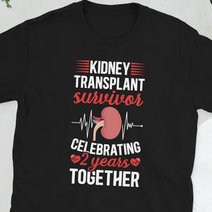 May include: Black t-shirt with white text that reads "Kidney Transplant Survivor Celebrating 2 years Together". A red kidney with blood vessels is shown with a heartbeat line above it. Two red hearts are on either side of the number 2.