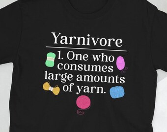 Op de afbeelding: Zwart T-shirt met witte tekst die "Yarnivore. 1. One who consumes large amounts of yarn." luidt. De tekst wordt omringd door kleurrijke garenbollen.