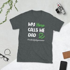 May include: Dark heather grey t-shirt with white text that reads "My Hero Calls Me Dad" with a green heart and ribbon. The text "#cerebralpalsyawareness" is below the main text.