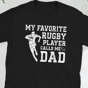 May include: Black t-shirt with white text that reads "My favourite rugby player calls me Dad". A white silhouette of a rugby player is holding a ball.