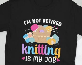 Op de afbeelding: Zwart T-shirt met de tekst "Ik ben niet met pensioen, breien is mijn werk" in wit. Het ontwerp bevat een mand met kleurrijke garenbollen, breinaalden en een spoel garen.