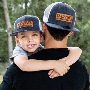 Könnte beinhalten: Ein Vater und sein Sohn tragen passende schwarze und graue Baseballkappen mit braunen Lederpatches, auf denen "DADVIBES" und "KIDVIBES" steht. Die Kappe des Vaters hat ein weißes Netzrücken.