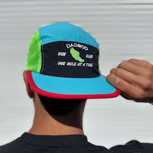 May include: A blue, black, and red Dadbod Run Club hat. The hat features a neon green mesh side panel and a red brim. The text "DADBOD RUN CLUB" and "ONE MILE AT A TIME" is embroidered on the front, with a green shoe graphic.