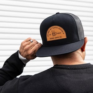 Pode incluir: Boné de caminhoneiro preto com um remendo de couro marrom que diz "Don't Tell But Always Shooting Up" com um logotipo de caveira e ossos cruzados e "Shroud Supply Co."