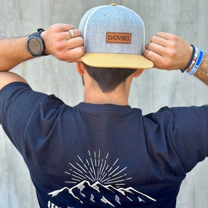 Könnte beinhalten: Ein marineblaues T-Shirt mit einem weißen Bergketten-Motiv und dem Text "Lead, Explore, Adventure" mit den Worten "Memories are waiting" über den Bergen. Der Text "Dadvibes" befindet sich unter den Bergen. Die Person trägt eine graue und beige Baseballkappe mit dem Text "Dadvibes" auf der Vorderseite.