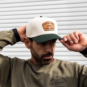 Pode incluir: Boné de beisebol creme com aba verde e um patch que diz "SUPPORT YOUR LOCAL GIRL DAD". O boné é usado por uma pessoa que veste um moletom verde.