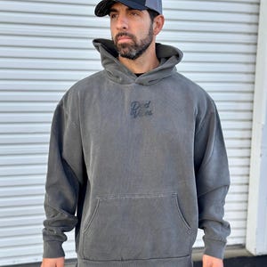 May include: A charcoal gray hooded sweatshirt with a black embroidered logo that says "Rad Vibes". The model is wearing a black and gray mesh baseball cap with a white logo that says "Rad Vibes".