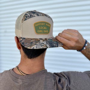 May include: A camouflage and beige baseball cap with a mesh back. The cap has a patch with the text "SUPPORT YOUR LOCAL GIRL DAD" in green and white.