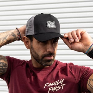 May include: A person wearing a black and gray mesh trucker hat with the text "Dad Vibes" on the front. The hat has a curved brim and a black strap. The person is also wearing a maroon t-shirt with the text "Family First" printed on the front.