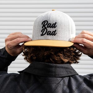 May include: A grey and yellow baseball cap with a black embroidered design that says "Rad Dad".