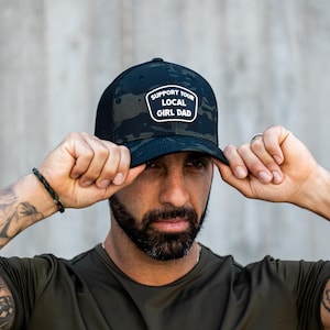May include: Black and camouflage baseball cap with the text "SUPPORT YOUR LOCAL GIRL DAD" on a white patch. The cap is worn by a person with a beard and tattoos.