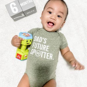 May include: A baby boy wearing a green bodysuit with the text "Dad's Future Spotter." He is holding a toy dumbbell and smiling. The baby is 6 months and 1 week old.