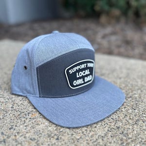 Può includere: Un cappello da baseball grigio e nero con una toppa che dice "Support Your Local Girl Dad".