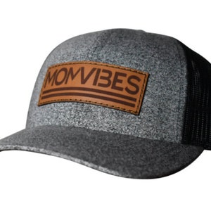 Puede incluir: Una gorra de camionero gris y negra con un parche de cuero marrón que dice "MOMVIBES" en letras marrones.