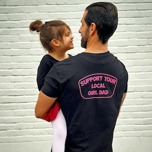 May include: A black t-shirt with a pink patch that says "Support Your Local Girl Dad". A young girl is being held by the person wearing the shirt.