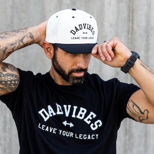 Könnte beinhalten: Weiß-schwarze Baseballkappe mit dem Text "DAD VIBES LEAVE YOUR LEGACY" in fetter, stilisierter Schrift. Die Kappe hat einen schwarzen Schirm und einen schwarzen Knopf oben. Das Bild zeigt auch ein schwarzes T-Shirt mit demselben Text.